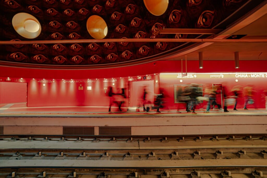 Passeggeri in transito sotto la cupola con occhi nella stazione Chiaia – Monte di Dio, area simbolica dell’Ade.