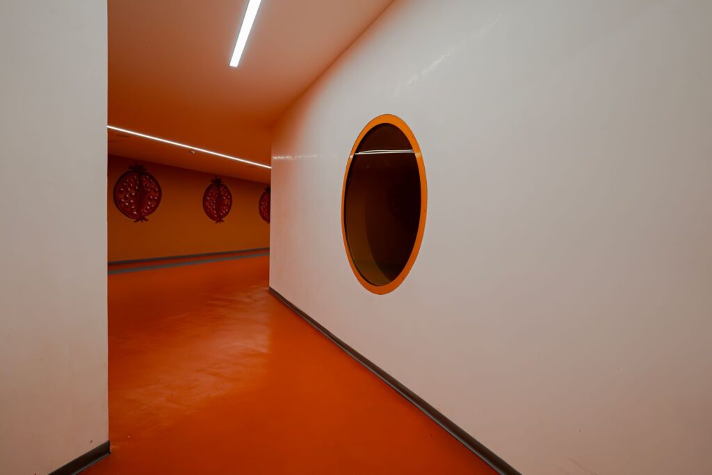 Corridoio arancione con finestra circolare e melograni, parte del percorso simbolico di Proserpina nella stazione.