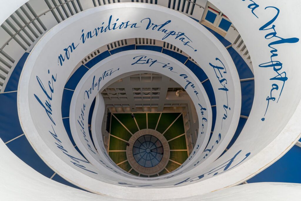 La spirale del cortile centrale della stazione Metropolitana di Chiaia – Monte di Dio, decorata con il nastro bianco e blu riportante il motto di Giulio Romano, elemento simbolico del percorso artistico ispirato all’estetica neoclassica e al dialogo tra luce e architettura.