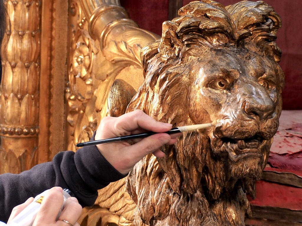 Dettaglio ravvicinato della pulitura manuale e laser sul muso del leone dorato che decora i braccioli del trono.