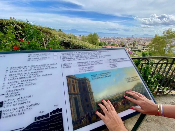 Una persona utilizza con le mani il pannello tattile dello skyline di Napoli presso il Belvedere di Capodimonte, con la città e il mare sullo sfondo.